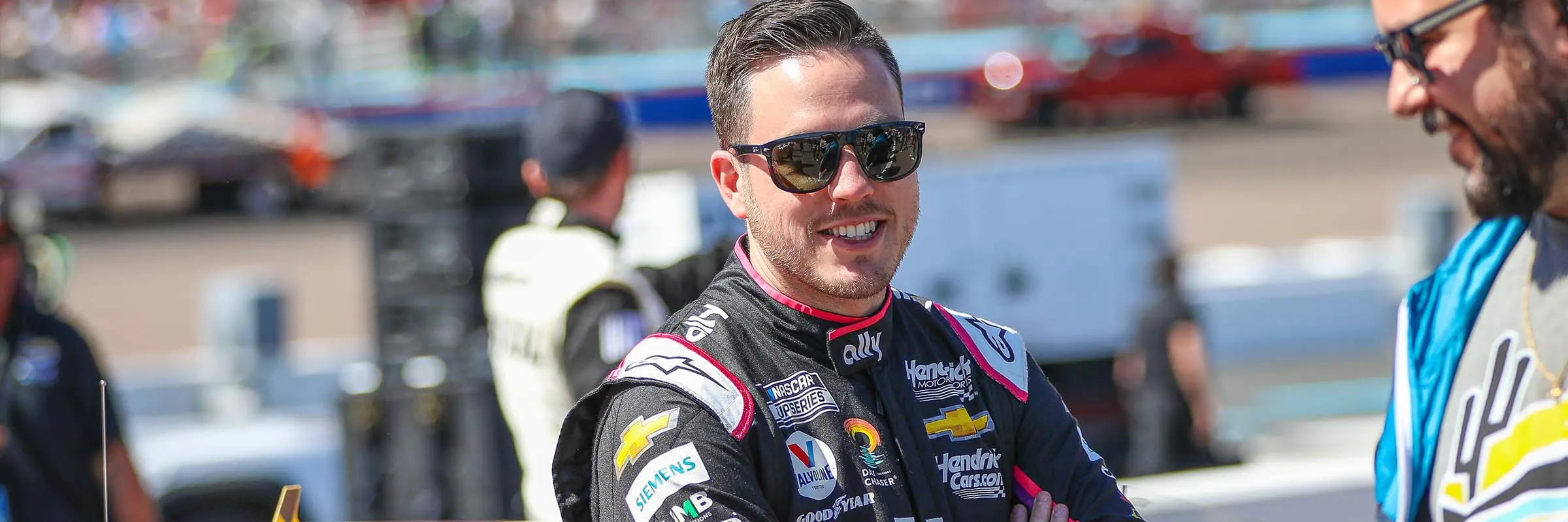 Alex Bowman leans against his NASCAR as smiles at a pit crew member 