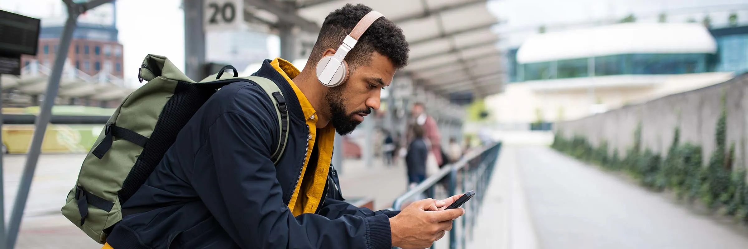 A young man is wearing headphones and looking at his phone while at a bus station.