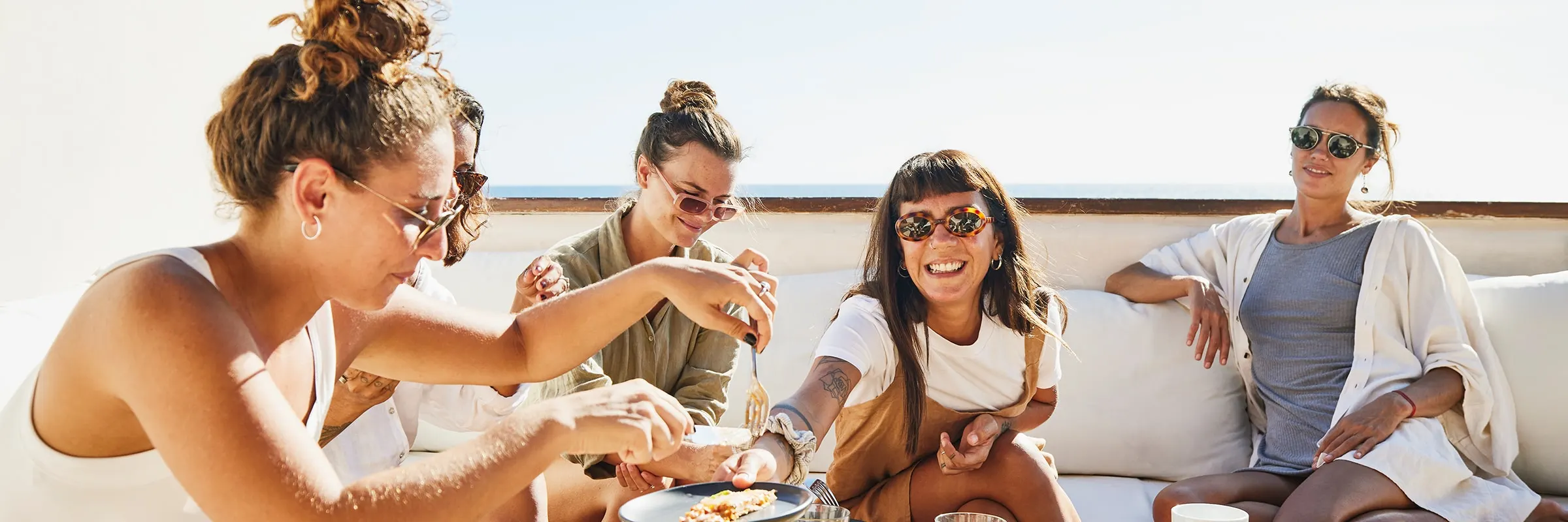 Group of young women sit outside on a balcony eating a meal together and laughing