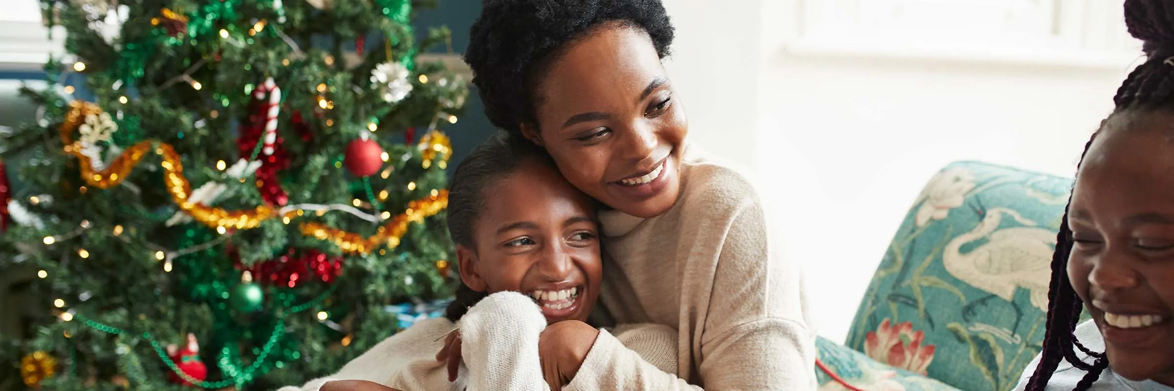 Smiling woman embracing a young girl while sitting by a Christmas tree.