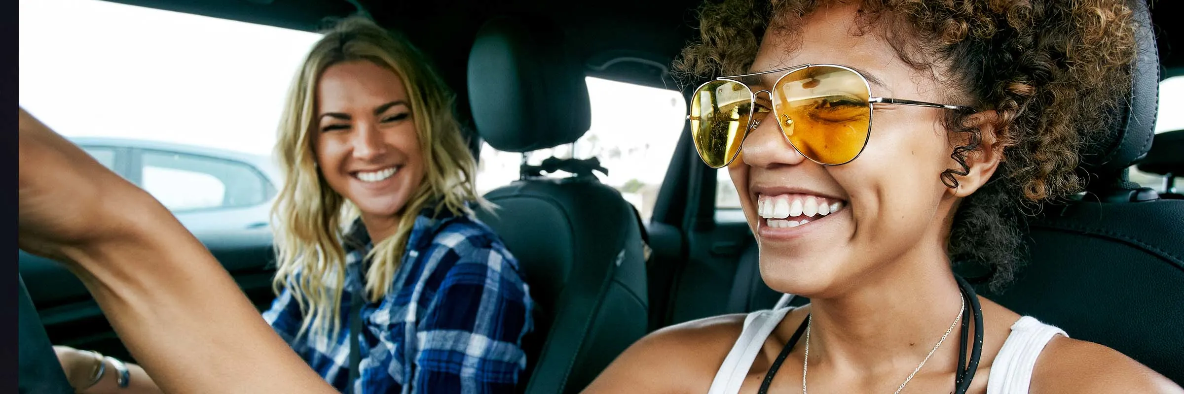 Two women, one with long blond hair and the other with brown curly hair sitting in car, wearing sunglasses, smiling.