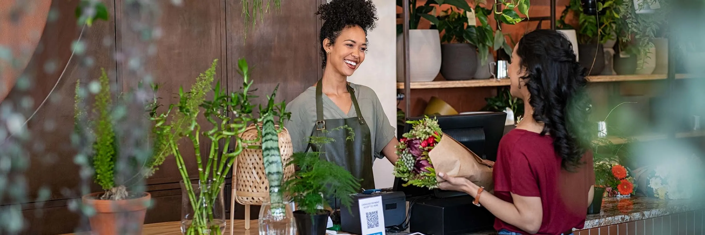 The owner of a flower shop is smiling while interacting with a customer at the cash register.