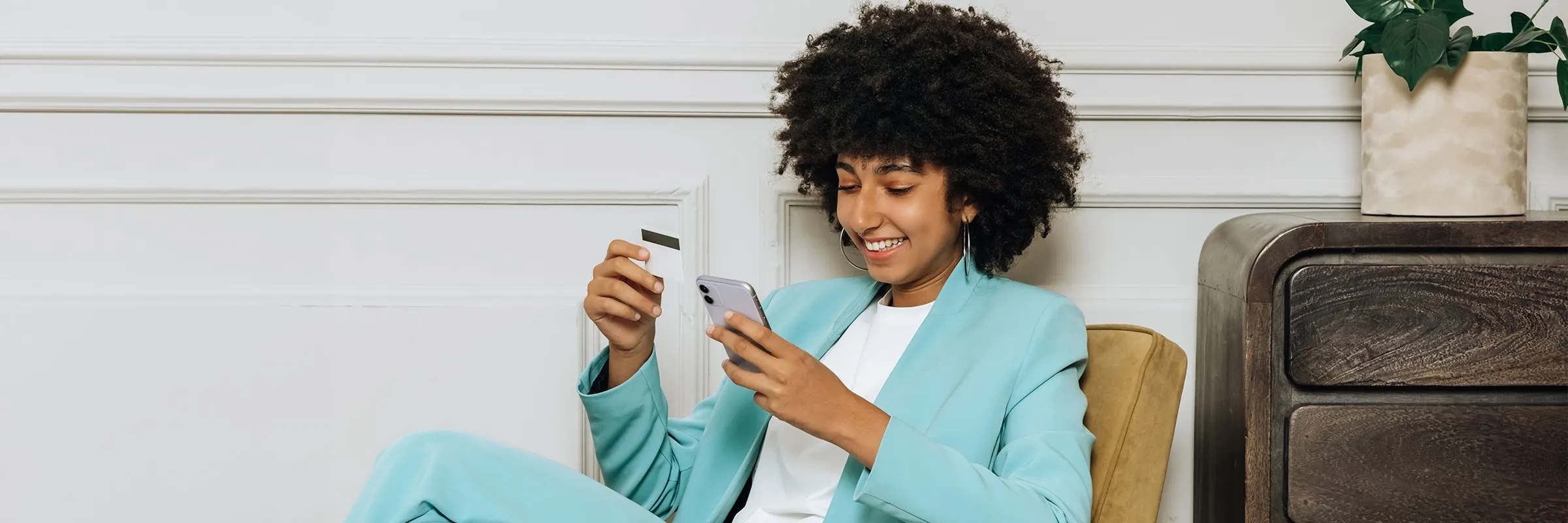 A woman is smiling while holding her phone and credit card in her hands to make an online purchase.