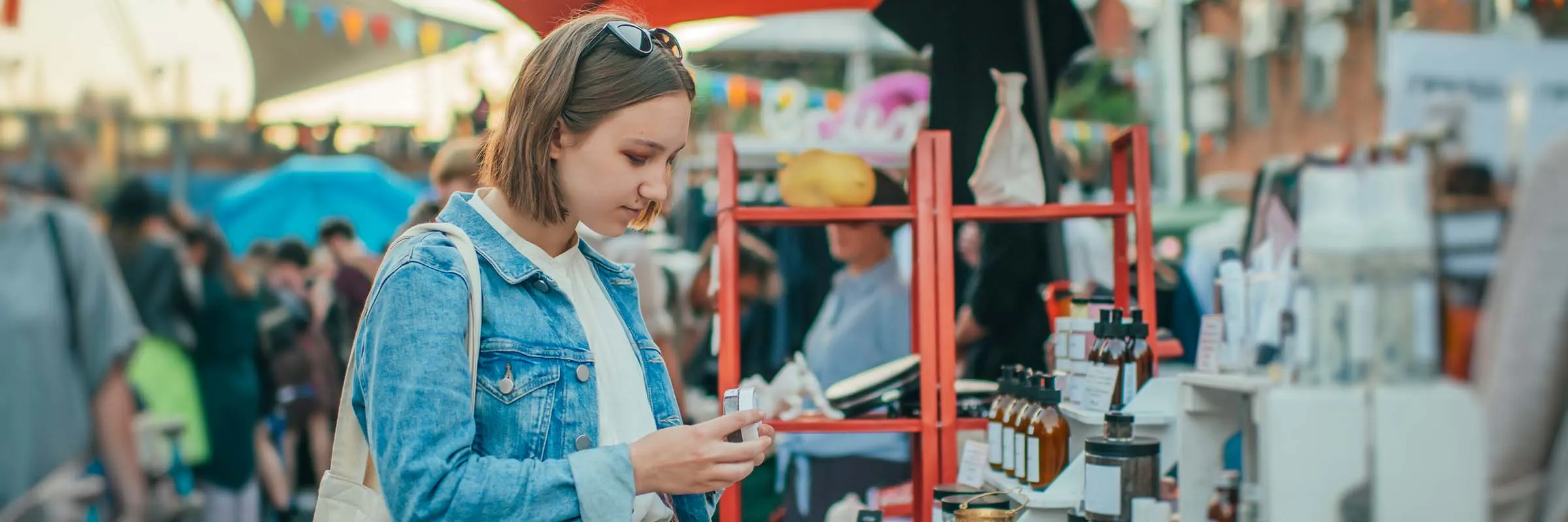 Young woman at a farmers market stands in front of a stall and looks at an item for sale.
