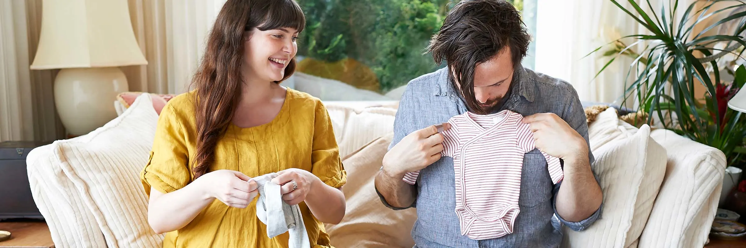 A pregnant woman and her partner are sitting on a couch and are looking at baby clothes.