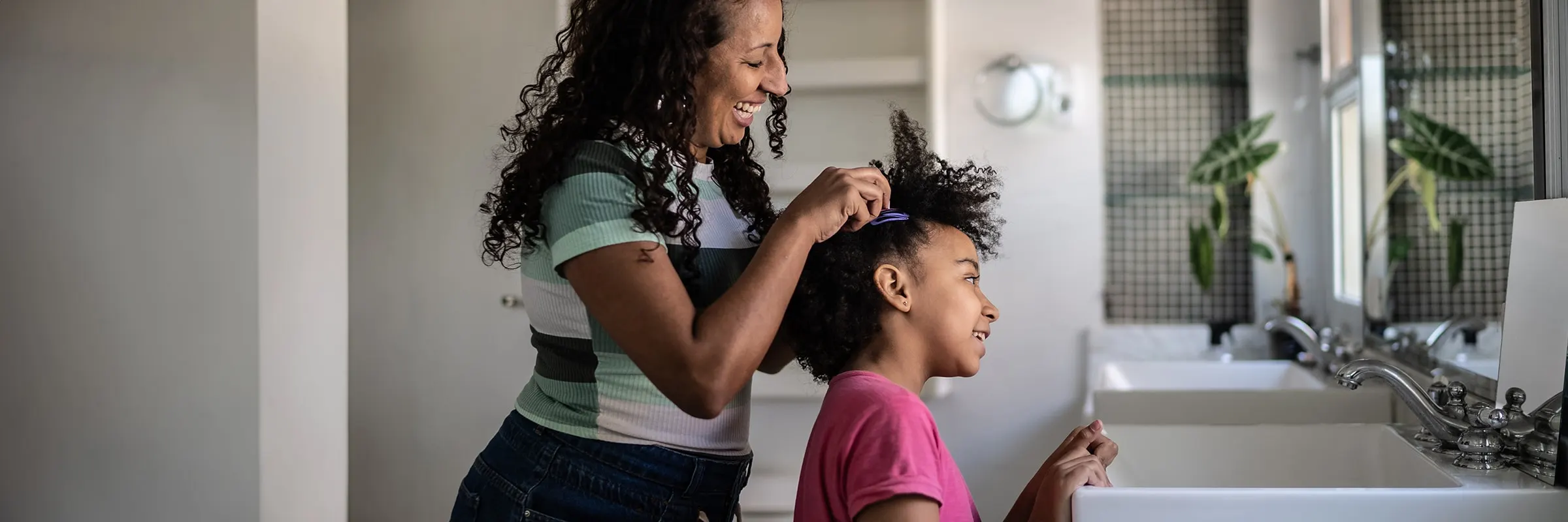 A mother smiles while combing her daughter's hair in a bathroom.