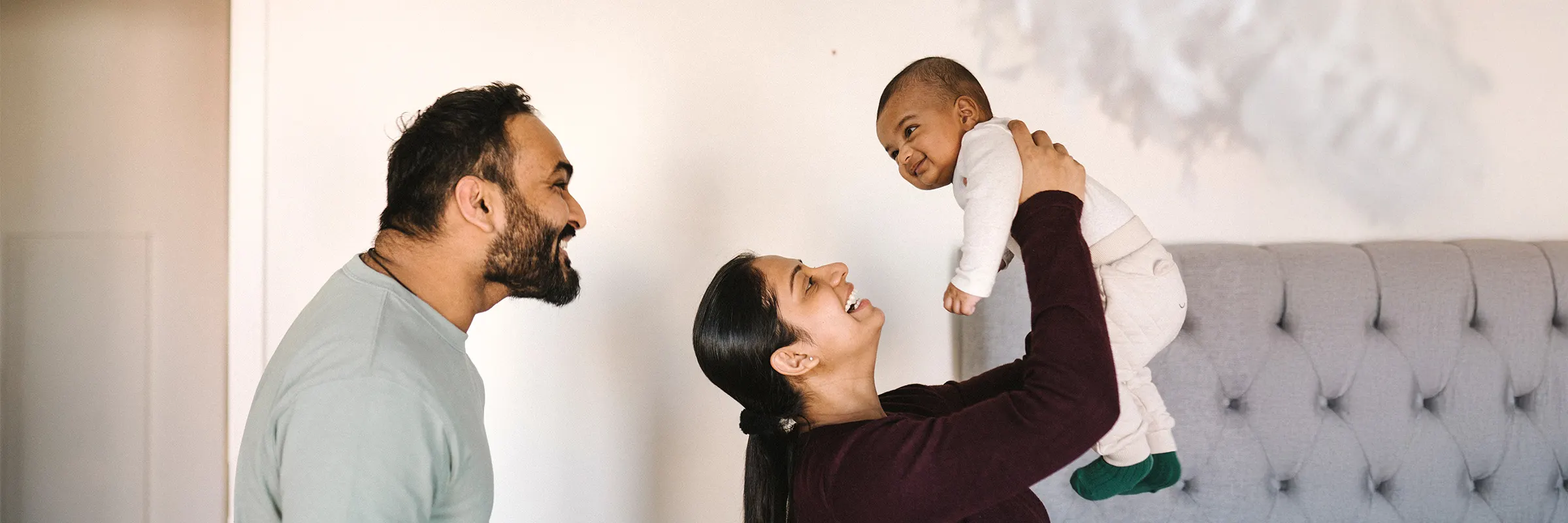 Two parents look on and smile at a small child. One person is holding the baby, the other is standing behind.