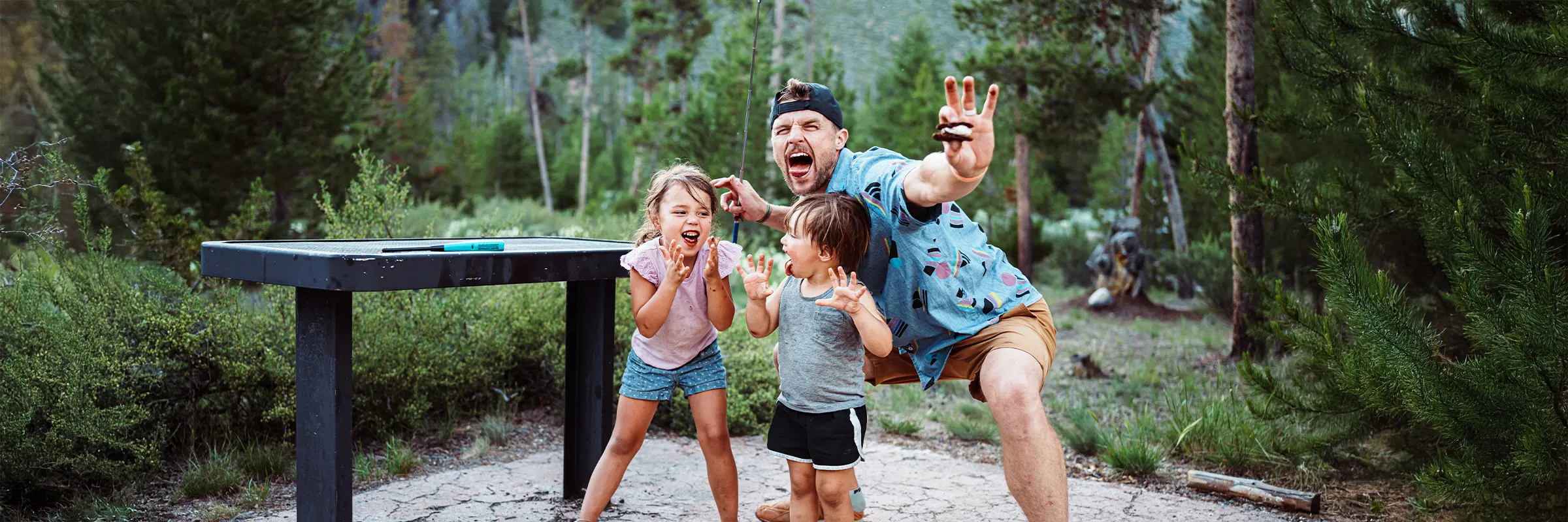 A father and two children at a campground making silly faces while eating smores.   