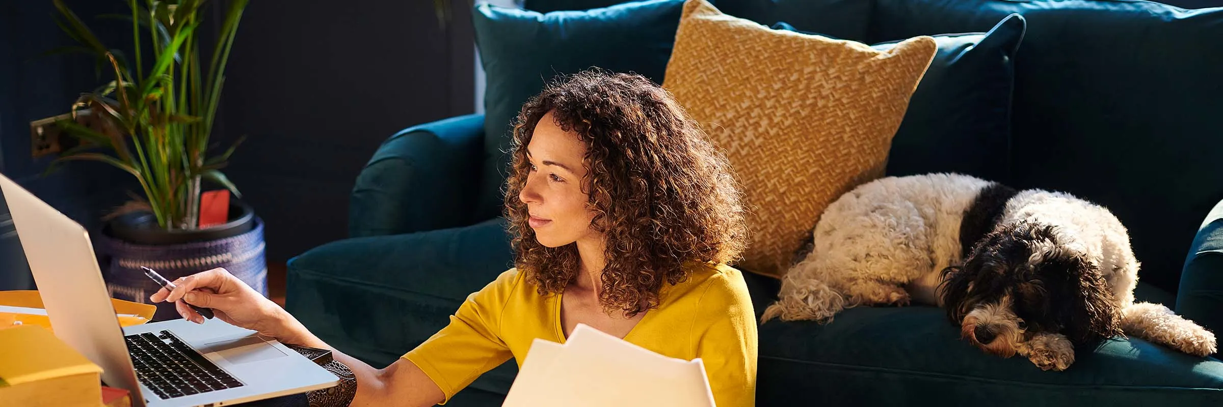Woman on her laptop while holding some paperwork. Dog on the couch behind her.