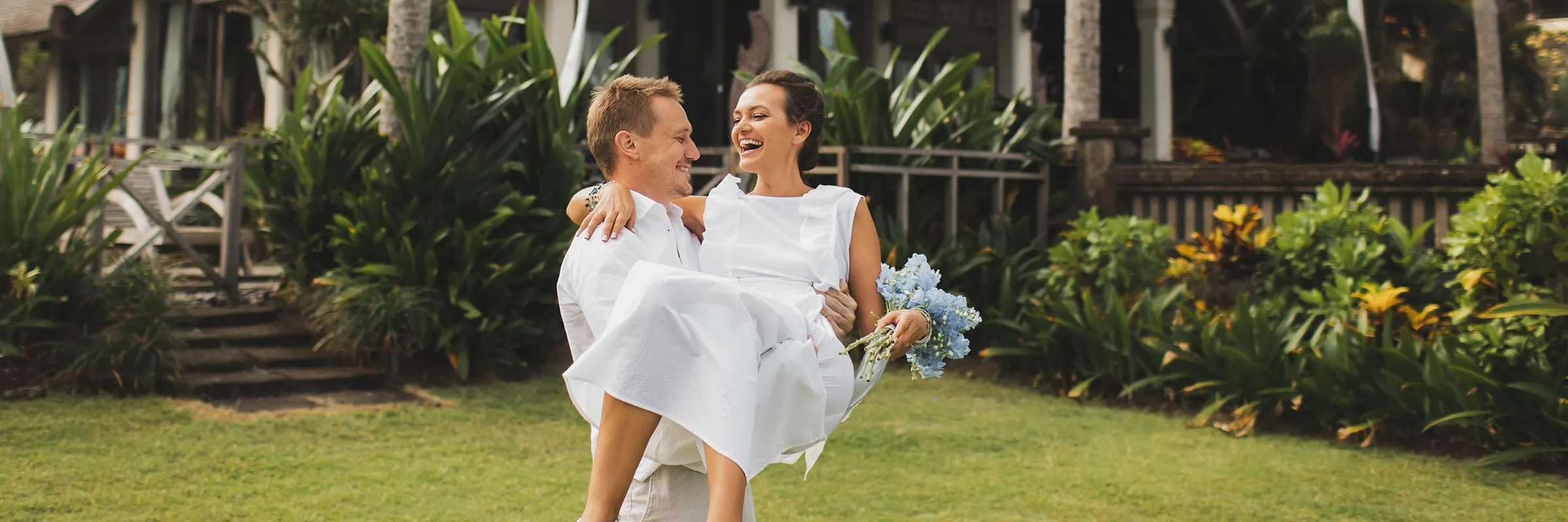  A newly married couple laughs on the lawn of a resort while the groom carries the bride.