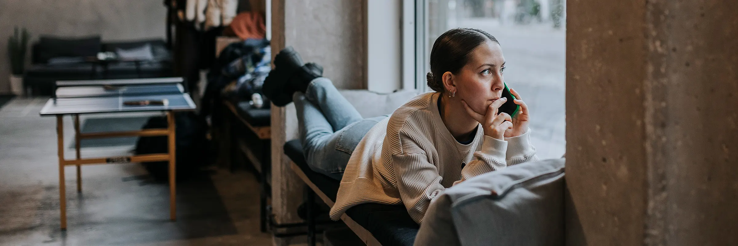 A woman lays out on a indoor bench and looks out the window while making a phone call. 