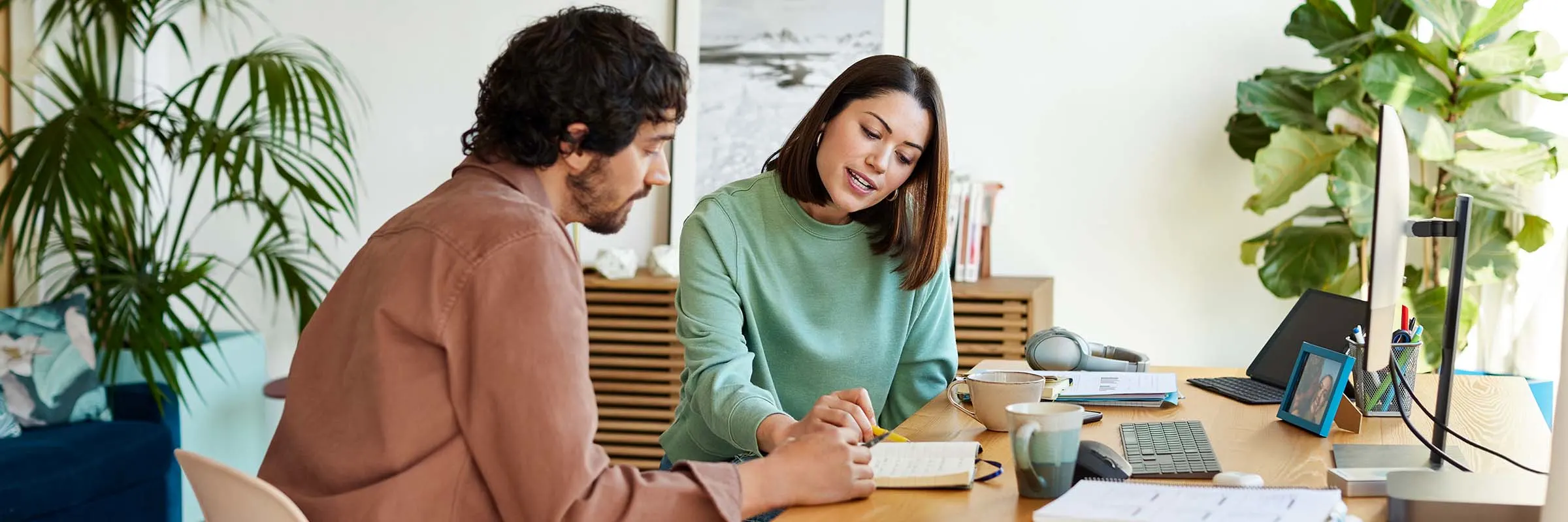 A man and woman are sitting at a desk and looking at a notebook together in a brightly lit home office.