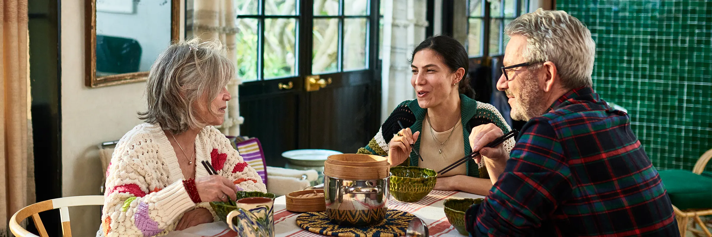 Two parents and their adult child sitting down for coffee at a dining room table.