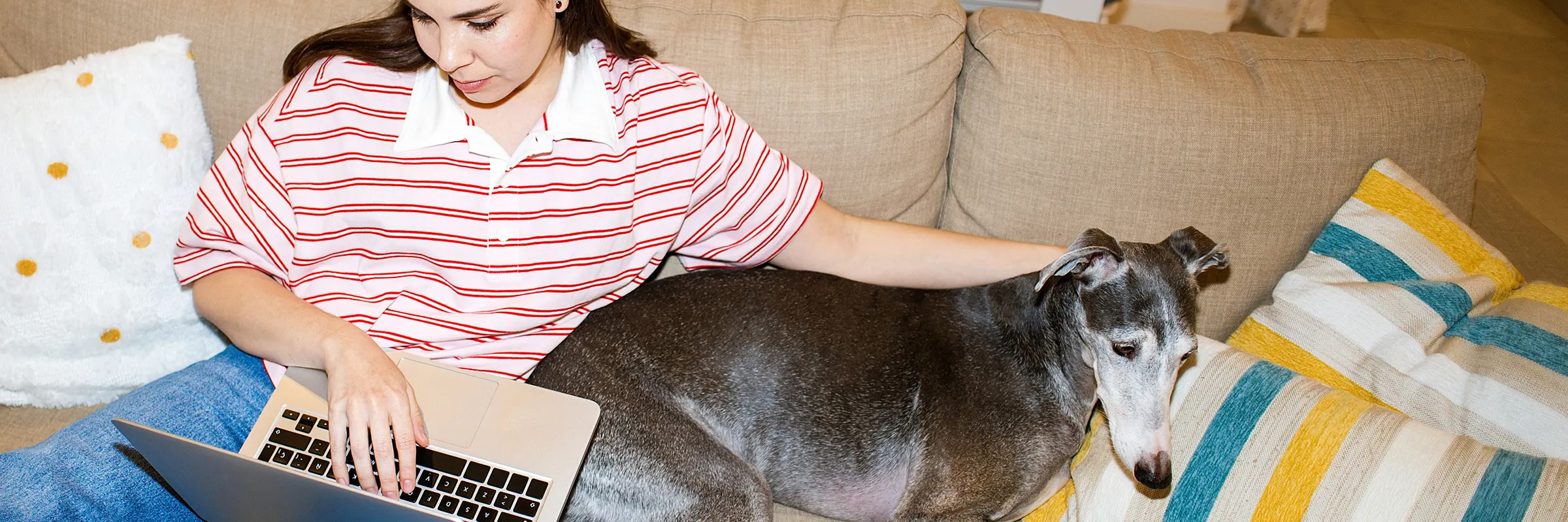 Woman with red hair on a couch looking at her laptop. Her dog sits next to her. 