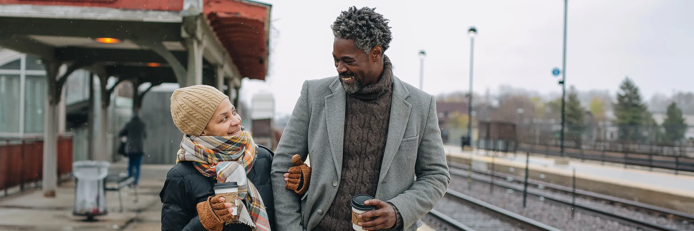 An older couple dressed in cold weather clothing walks down a train platform with their arms linked and coffee in their hands.