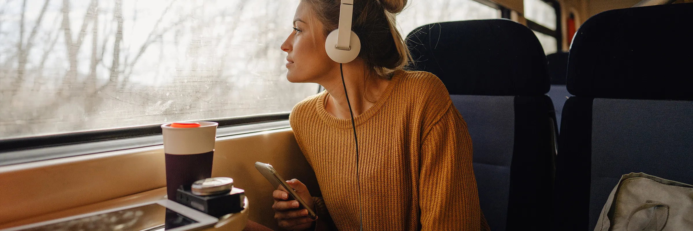 A young woman is riding on a train, looking through the window and listening with headphones to her mobile phone.