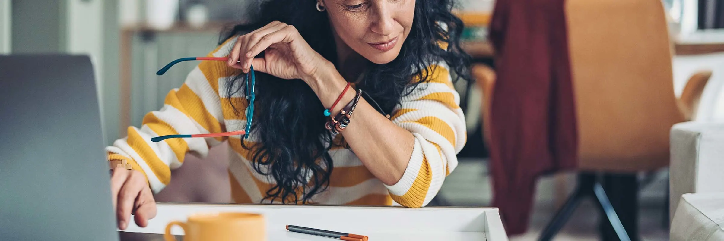 A woman looks at a piece of paper on her desk. She holds her glasses in one hand.