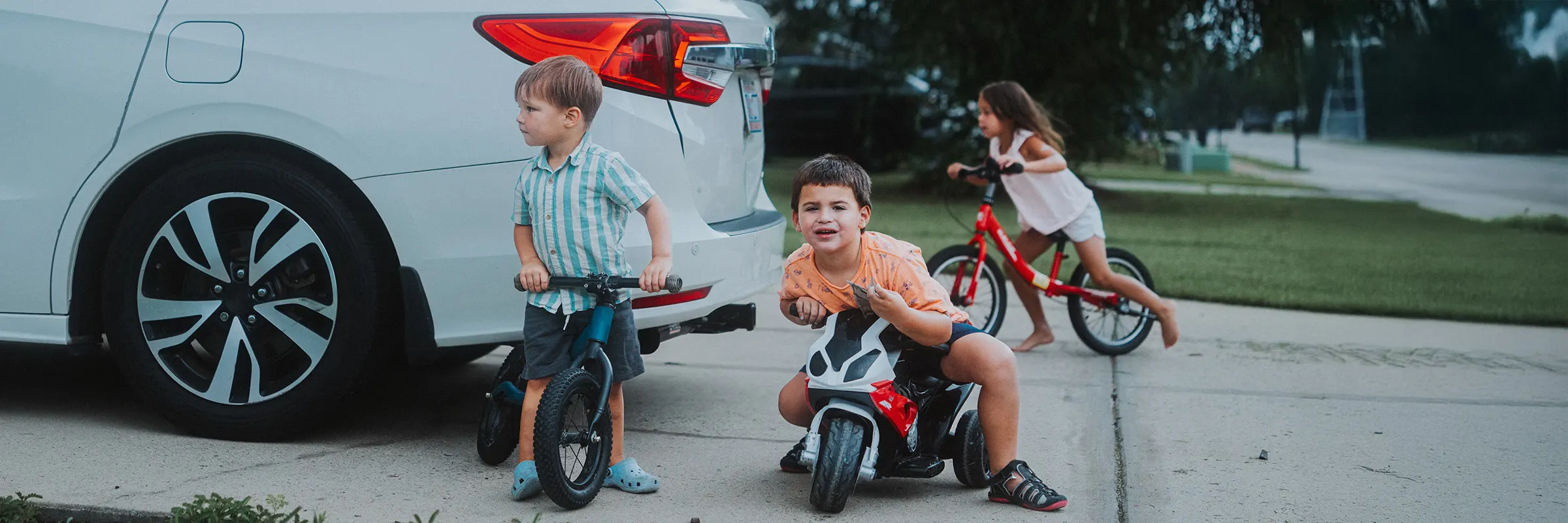 three children ride bikes and play around a van in a driveway