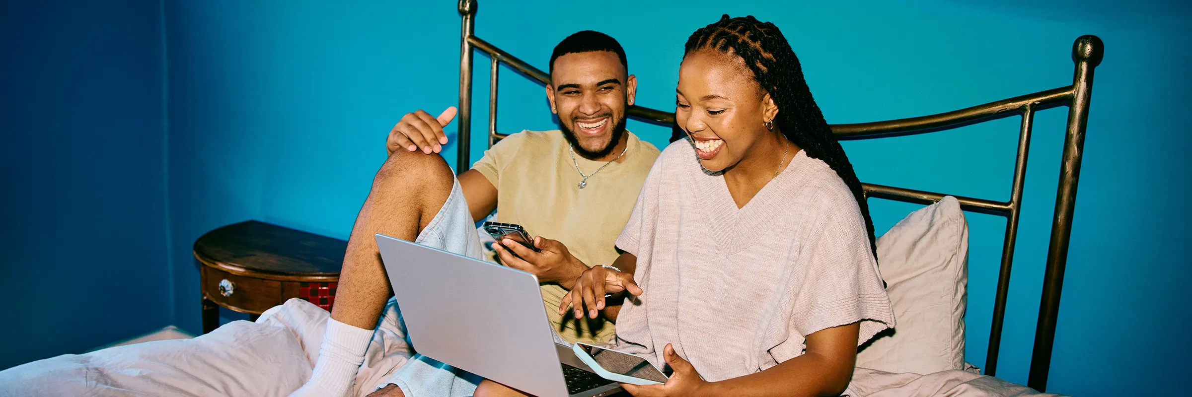 Wide shot of a cheerful young couple sitting on a bed with a laptop in a bright blue room