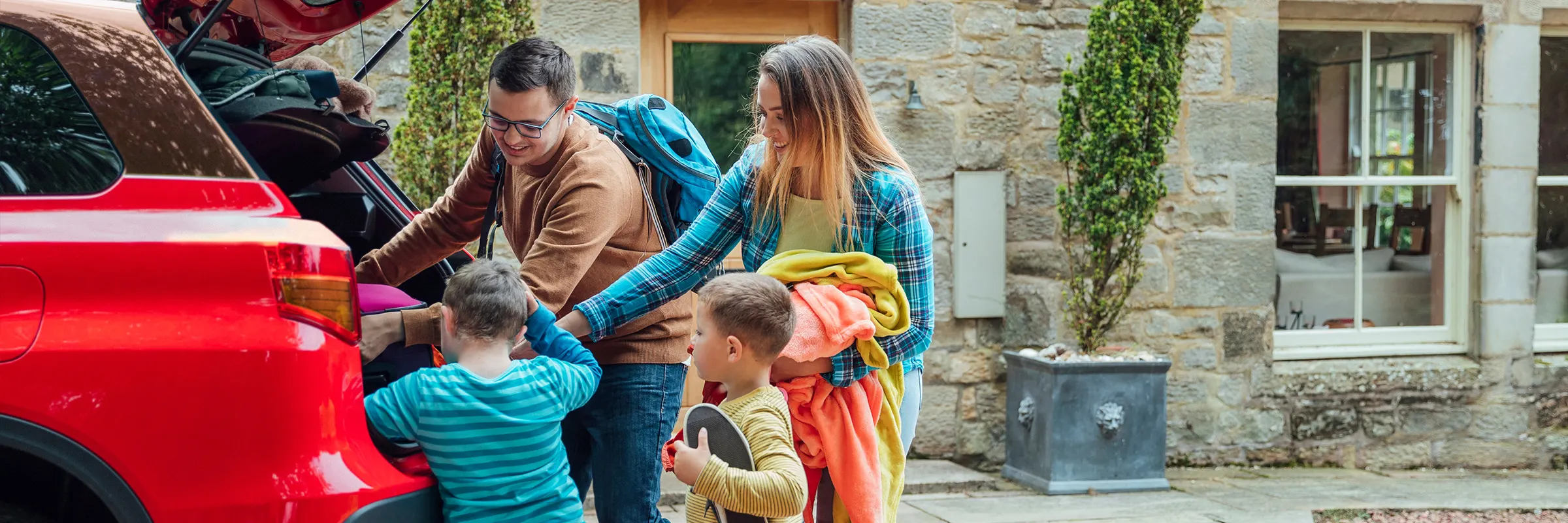 A man and woman help their two young children pack the car for a road trip. 