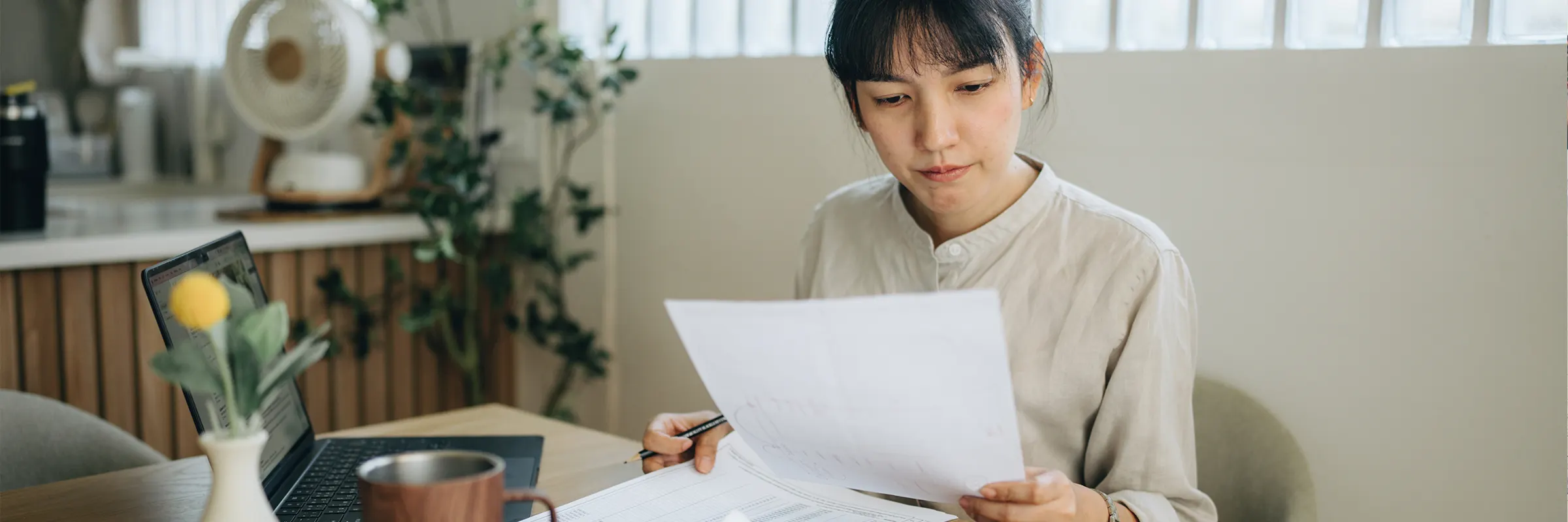 A person sits at their kitchen table looking at a document while their laptop is open.