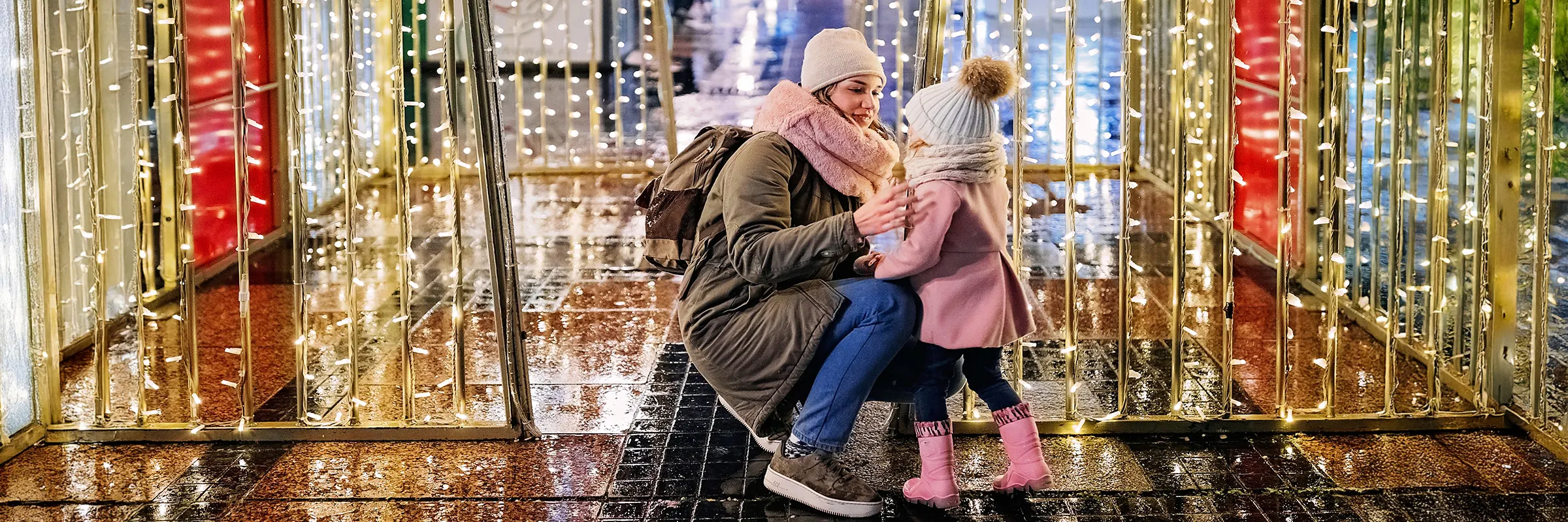 While standing in front of holiday lights outside, a mother kneels down to help her child with winter clothing 