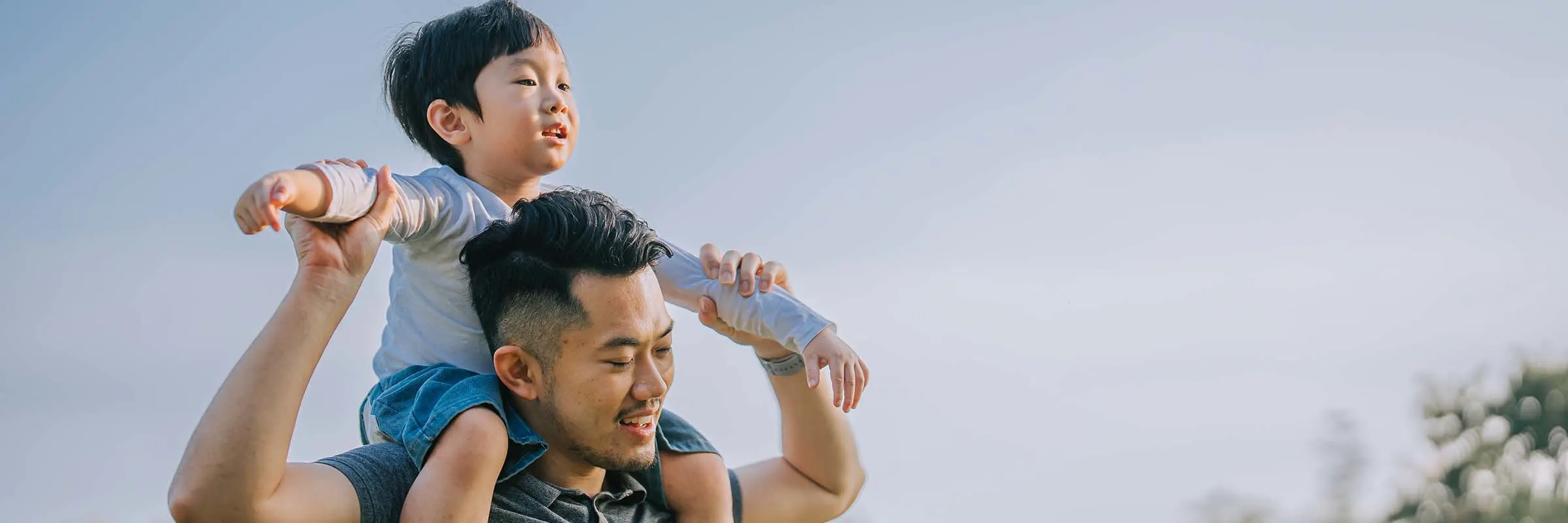 Man holding his child on his shoulders, both smiling outside.