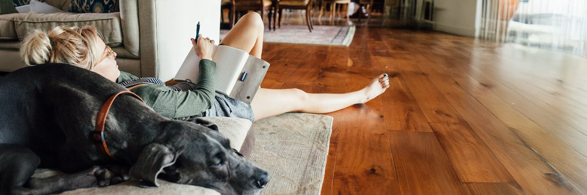 Young adult lays on the floor with their back leaning against a sleeping Great Dane.