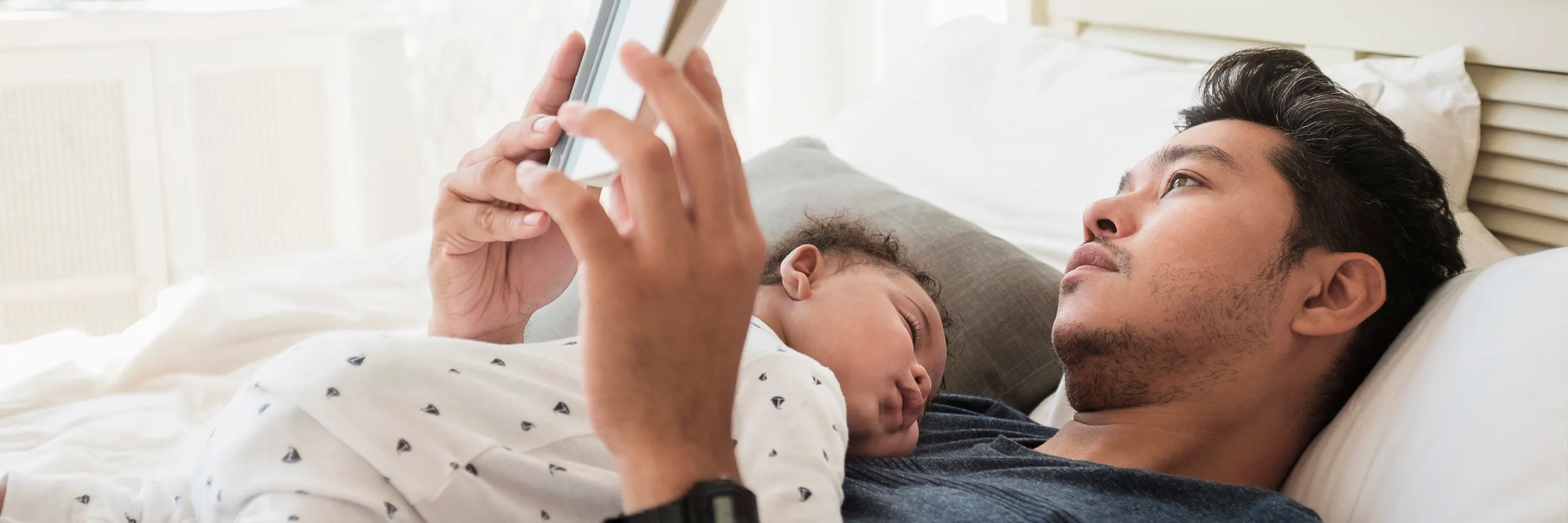 A father reads a book in bed while his baby sleeps on his chest. 