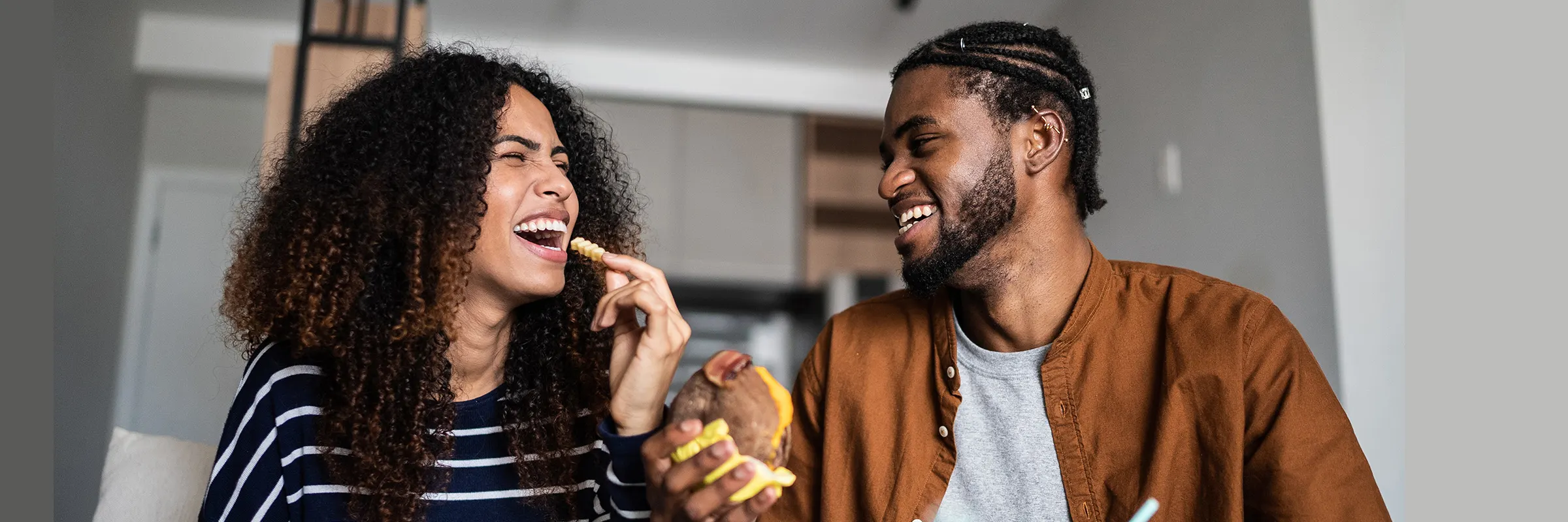 A young couple laughing while eating fast food on the couch. 