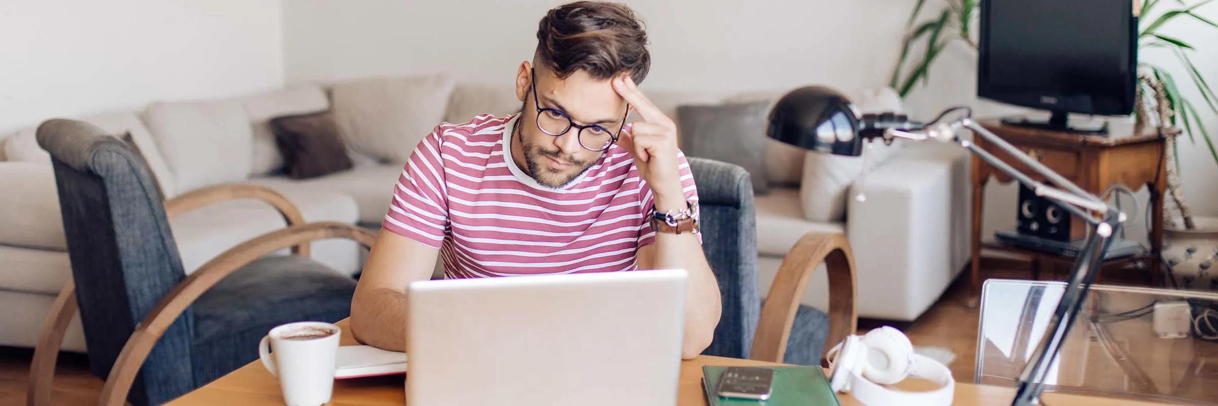 A young man is working from home at a table in his living room. He is wearing glasses and looking at his laptop.
