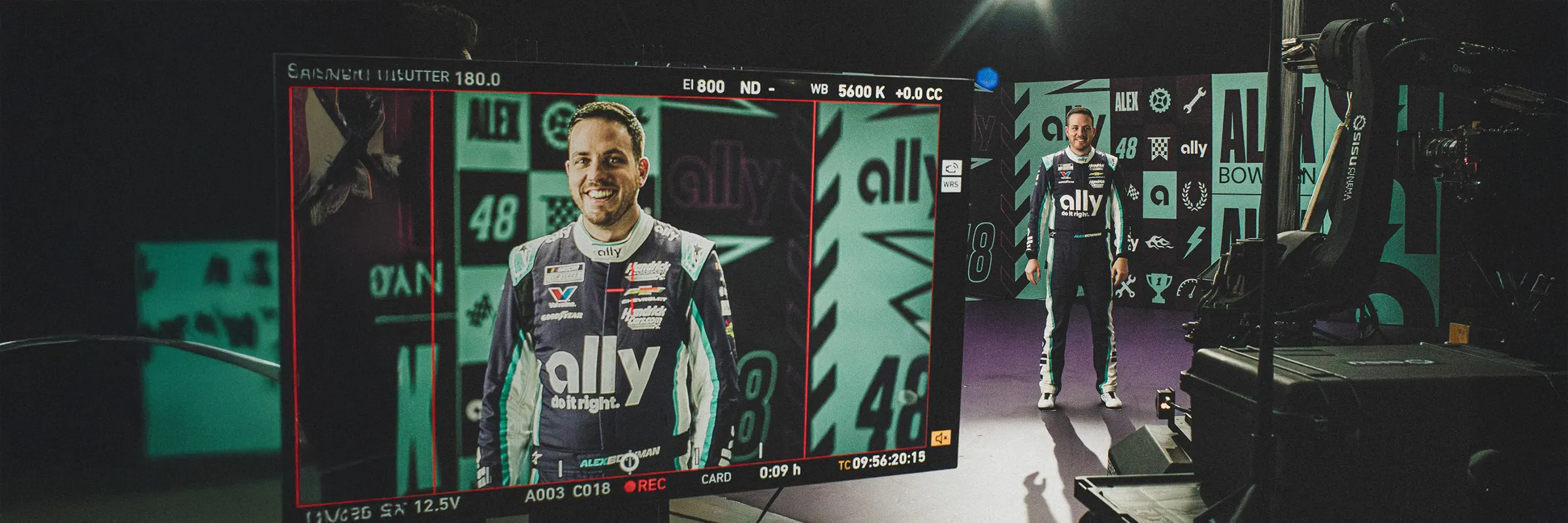 Alex Bowman, driver of the #48 Ally racecar, stands in the background while his image is on monitor in foreground.