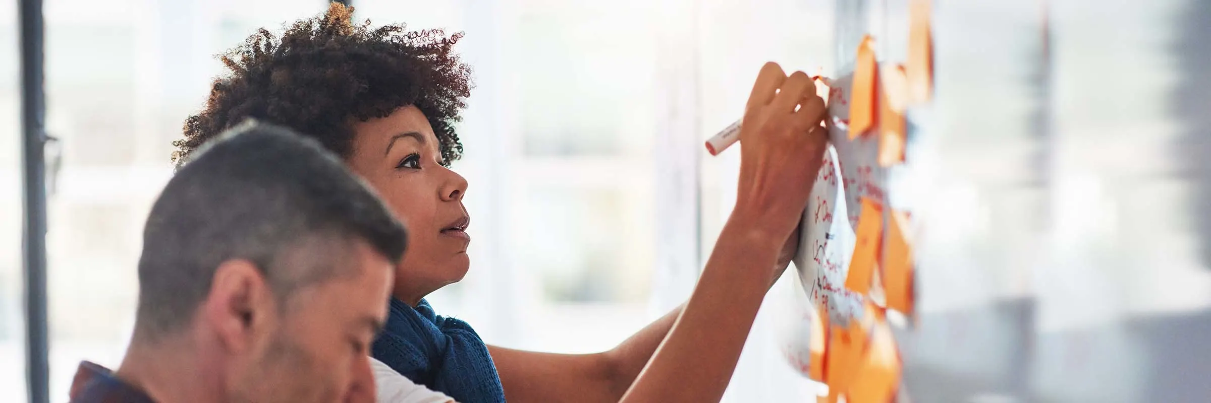 A woman takes notes on a whiteboard while a man looks at sticky notes placed on the whiteboard.
