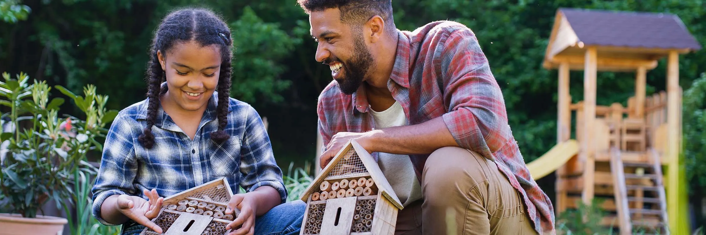 A young man is in a backyard with his younger sister. They are each holding bug hotels and smiling.
