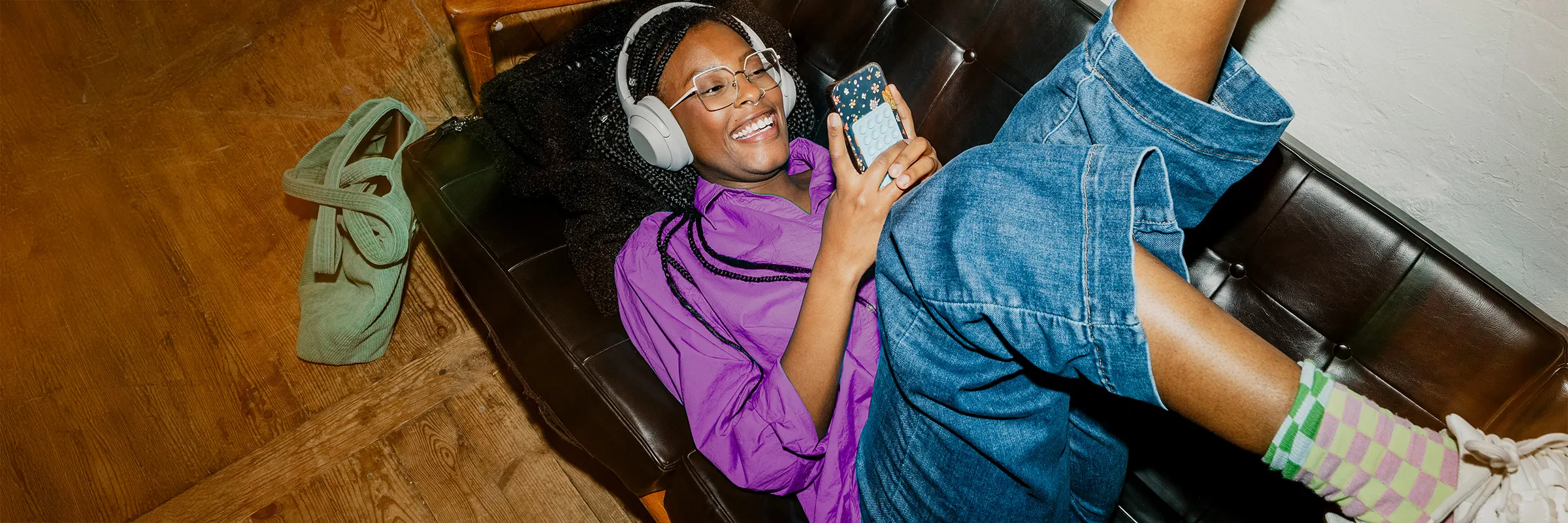 Young woman scrolling on her mobile device with headphones while lying on the couch. 