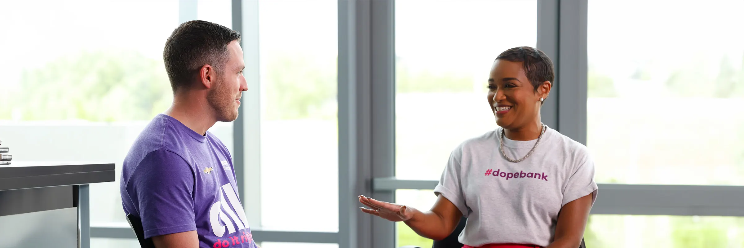 Hendrick racecar driver, Alex Bowman, sits beside Ally’s Jack Howard. They are smiling at each other while talking. 
