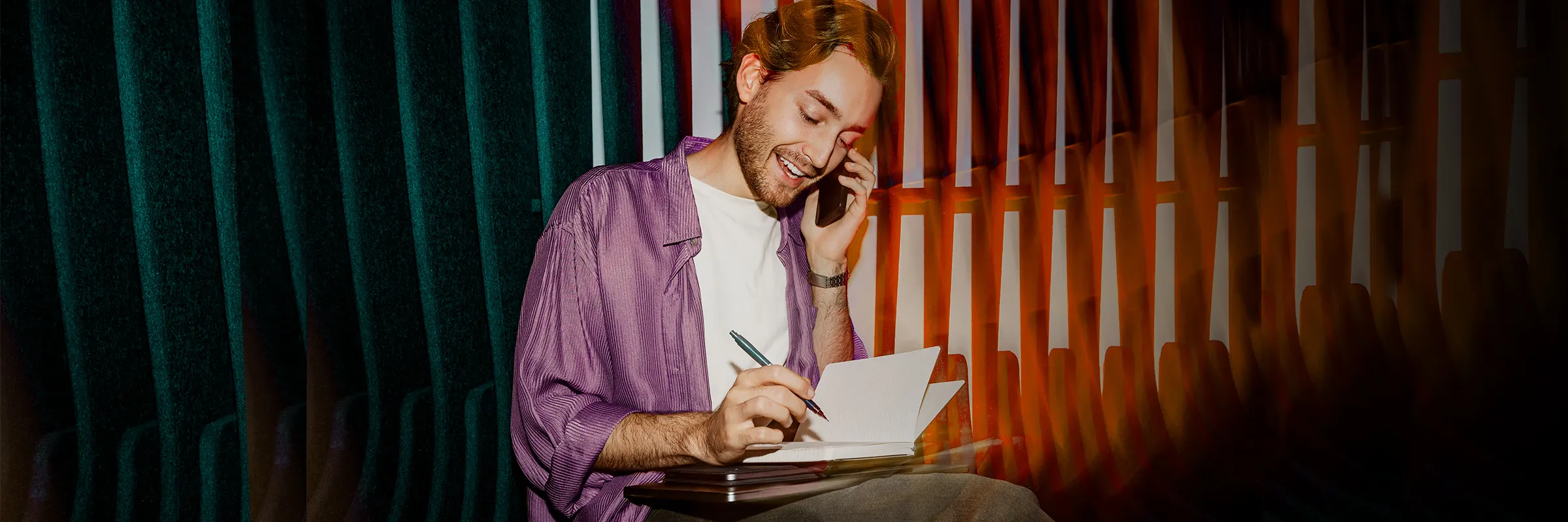 Young man sitting in a chair taking notes while on a phone call. 