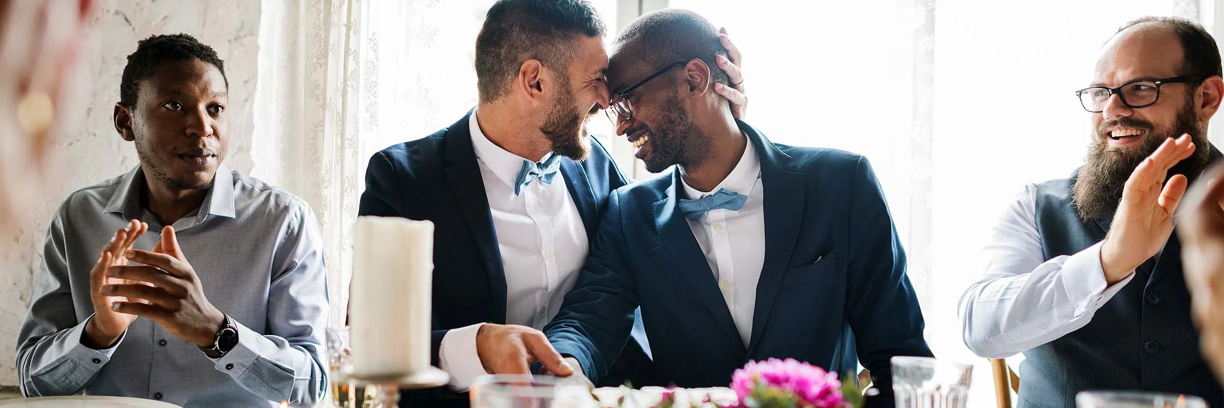 Two grooms are embracing while seated at the head table at their wedding.