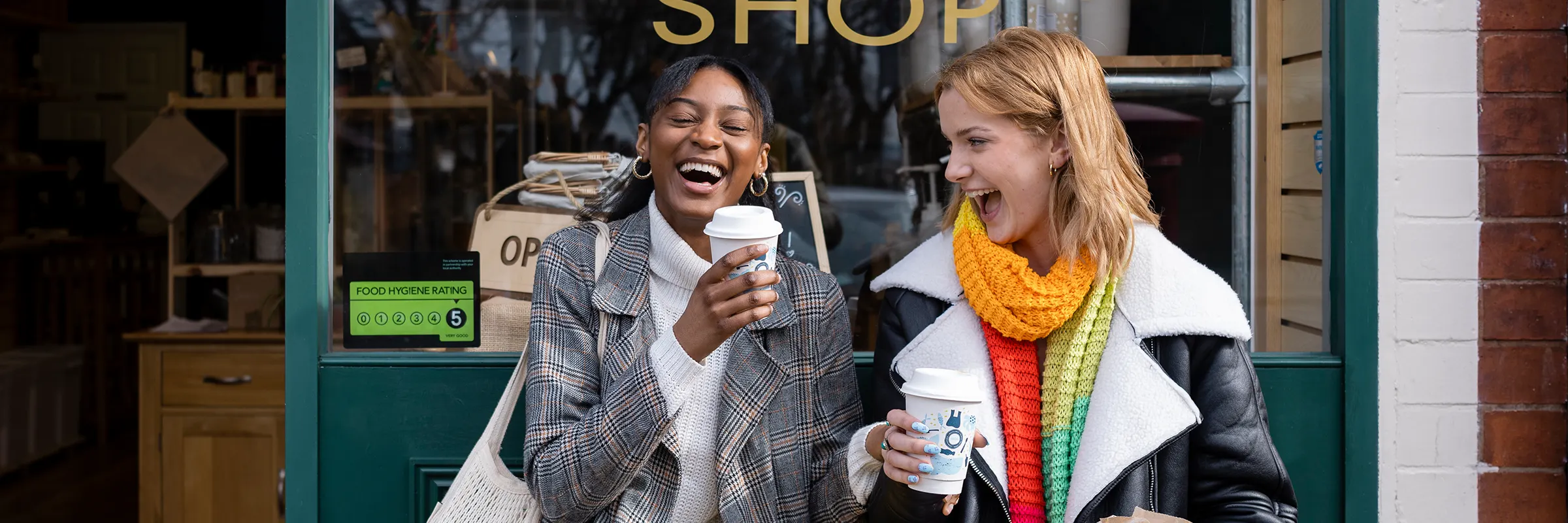 Two friends sitting in front of a shop laughing while holding coffee.