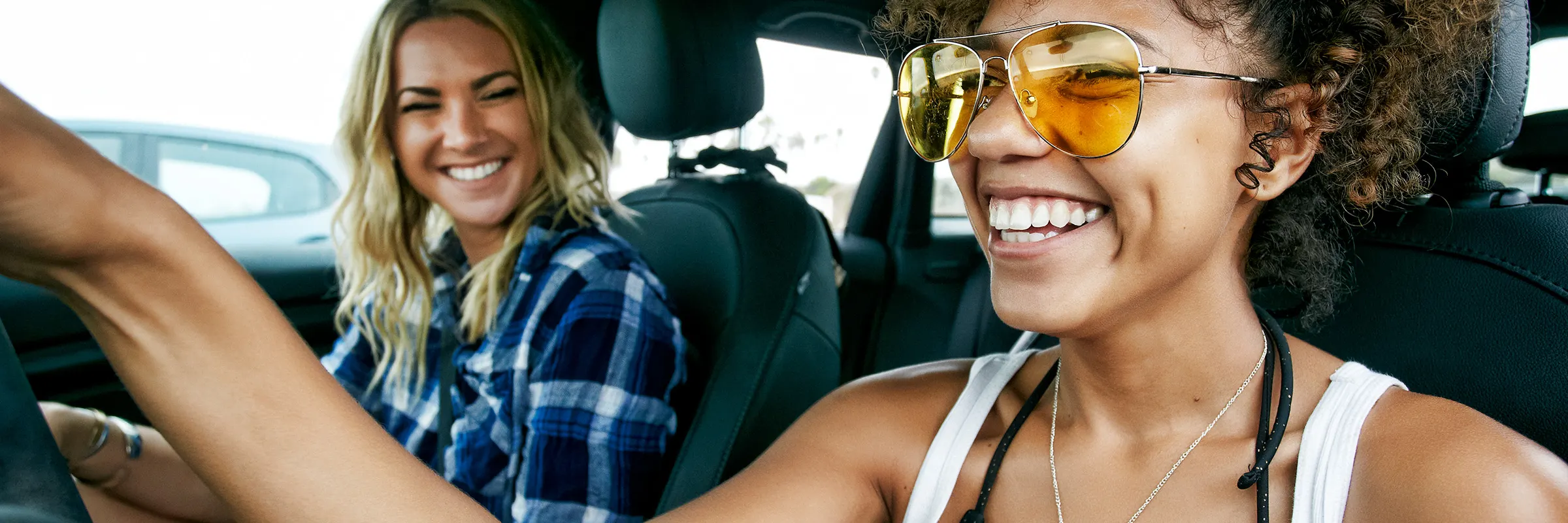 Two women, one with long blond hair and the other with brown curly hair sitting in car, wearing sunglasses, smiling.