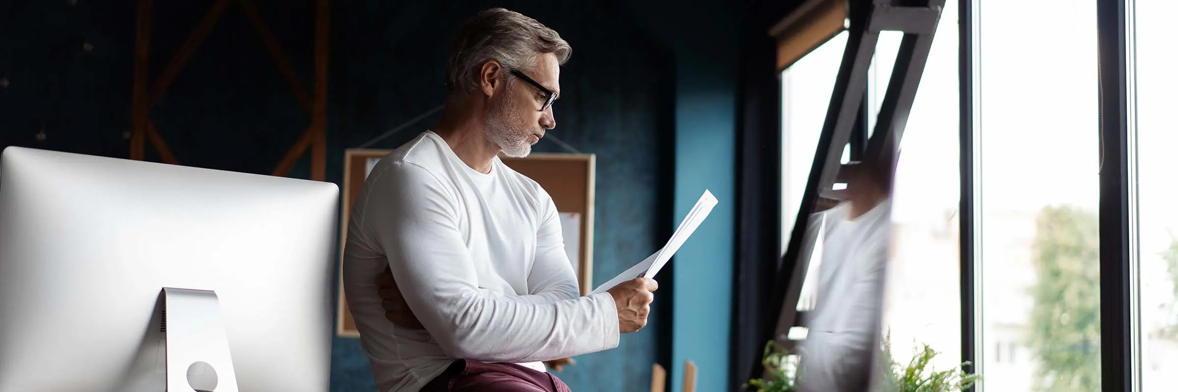 A man sits on a desk in his home office and reads a piece of paper.