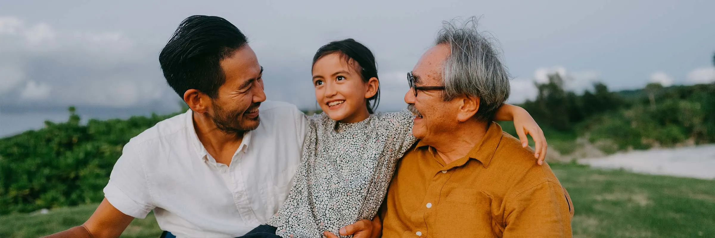  Three generation family, an older man with his son and grandchild, sit outside smiling at each other