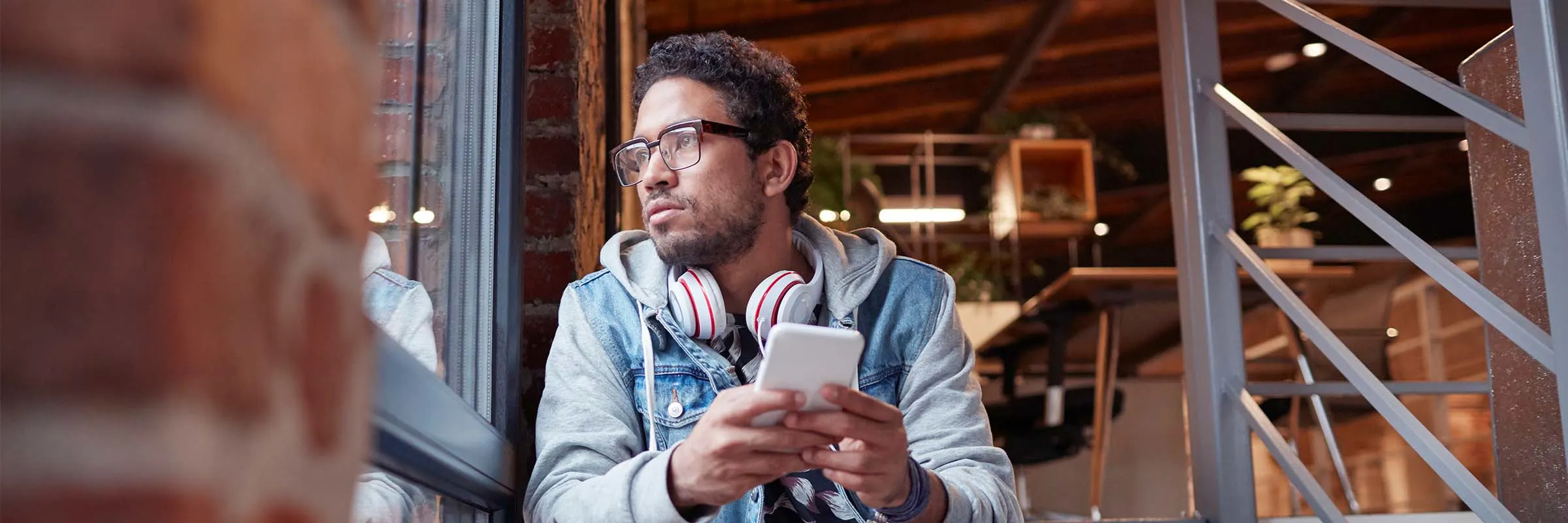 Young man sitting on the steps in an office looking out the window while holding a phone. 