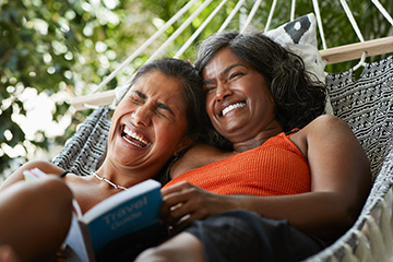 Two woman lounging in a hammock while laughing and enjoying a book.