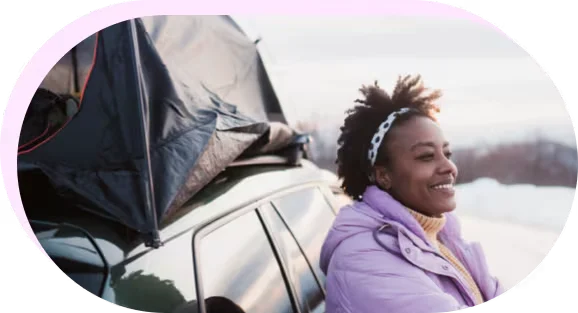 a smiling woman leaning against her vehicle gazing at the snow.