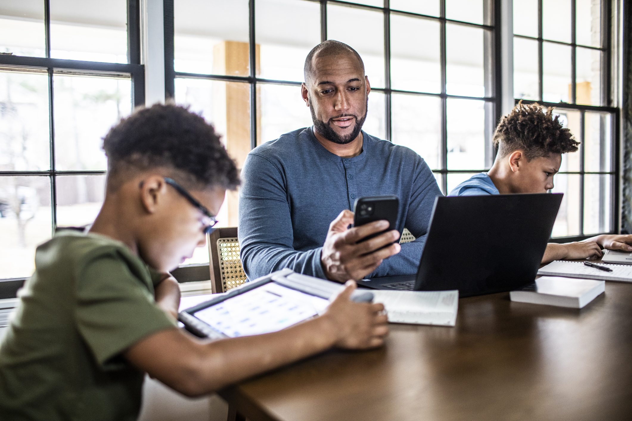 Father and two young sons sitting at a table with technology. The father is looking at his smartphone with his laptop open in front of him.