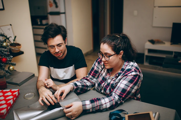 Couple sits at a table while wrapping gifts together.
