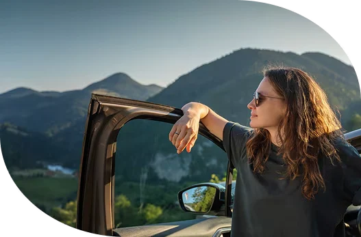 a woman standing inside the driver’s side door of her vehicle gazing at the mountains.