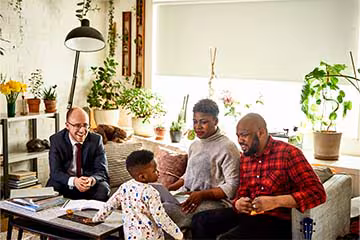  Two parents sitting on their couch with a laptop and their financial advisor. Their toddler son is looking up at the father and there are many plants in the background.