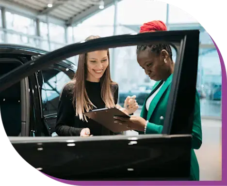 Two women standing behind an open car door in a dealership looking at a clipboard.